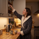 Woman in a charcoal robe reaching for a mug in a warm, cream kitchen.
